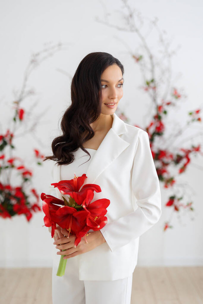 Bride in a white trouser suit posing against the background of an unusual composition of fresh flowers poinsetia for an chamber wedding ceremony. Hold bouquet of red amaryllis. - Фото, зображення