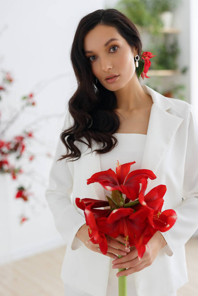 Fashion portrait of young woman poising in white trouser suit. Wedding trends and concept. Holding bouquet of red amaryllis. - Fotografie, Obrázek