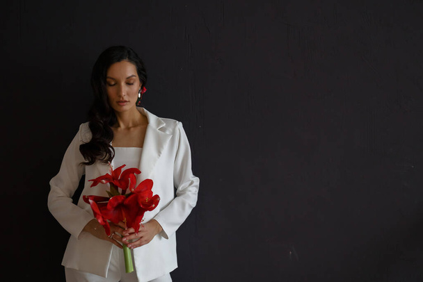 Fashion portrait of young woman poising in white trouser suit. Wedding trends and concept. Holding bouquet of red amaryllis. Dark background - Fotoğraf, Görsel