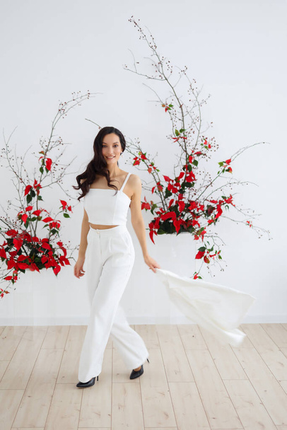 Happy Fashionable bride in a white trouser suit. Ceremony arch of red poinsettia. Wedding in winter season. - Foto, Imagen