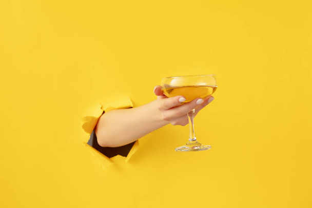 Female hand with cocktail glass breaks through yellow paper background. - Foto, afbeelding