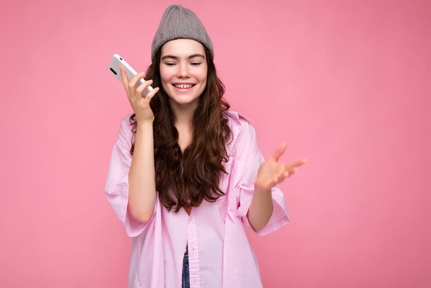 Pozitív mosolygós vonzó fiatal barna nő visel stílusos rózsaszín ing és szürke kalap elszigetelt rózsaszín háttér gazdaság a kezében, és a mobiltelefon kommunikáció és hangüzenet rögzítése - Fotó, kép