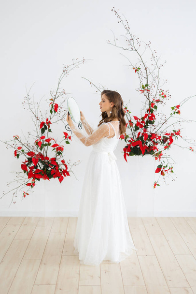 Happy bride in a white wedding dress looks at herself in the mirror. Red flowers on the background. - Фото, изображение