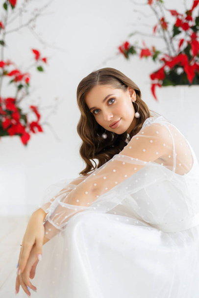 Bride in dress sitting on floor. Wedding arch and decor with red poinsettia flowers. Indoor bride morning. - Photo, image