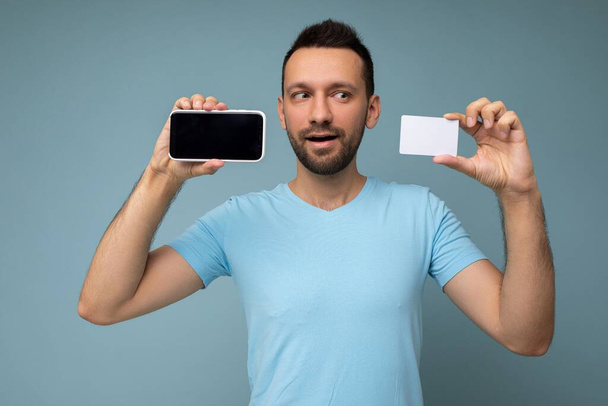Фото красивого молодого брюнетки с бородой в повседневной синей футболке на синем фоне, держащего и использующего мобильный телефон и кредитную карту, делающего платежи, глядя в сторону - Фото, изображение