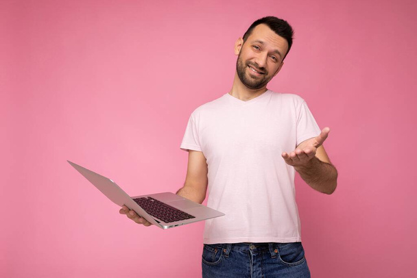 Fešák legrační a usměvavý brunet muž drží notebook a ukazuje ruku při pohledu na kameru v tričku na izolovaném růžovém pozadí - Fotografie, Obrázek