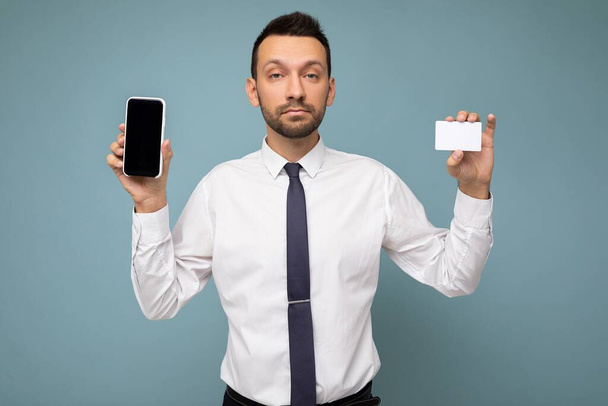 Photo of handsome good looking brunet man with beard wearing casual white shirt and tie isolated on blue background with empty space holding in hand and showing mobile phone with empty screen for - Фото, зображення