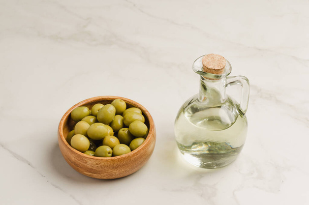 Bottle of olive oil and olives in a wooden bowl. Olive oil bottle and olives on white stone table - Valokuva, kuva