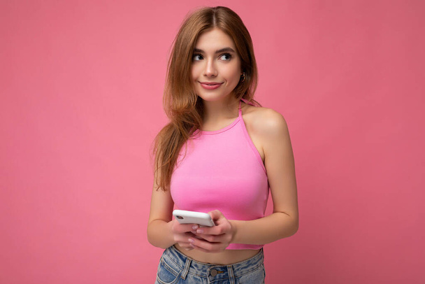 Photo of sexy good looking young blonde woman wearing pink top poising isolated on pink background with empty space holding in hand and using mobile phone messaging sms looking to the side - Фото, изображение