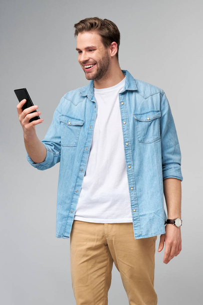 Smiling young man wearing jeans shirt taking selfie photo on smartphone or making video call standing over grey studio background - Valokuva, kuva