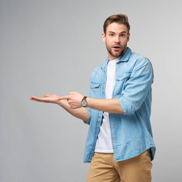 Happy young handsome man in jeans shirt pointing away standing against grey background - Photo, image