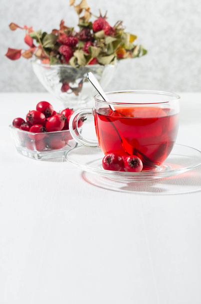 Natural red herbal tea in transparent cup with hawthorn berry, rosehip, dry leaves in morning sunlight in soft light white interior, copy space, vertical. - Фото, зображення
