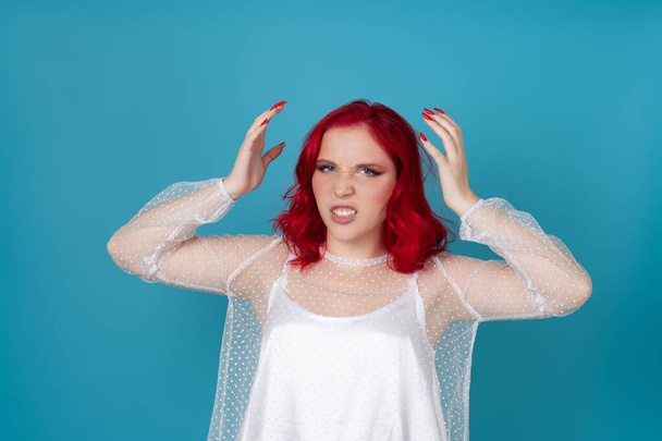 a frustrated, angry woman in a white silk dress and with red hair clutches her head with her hands from resentment and insult , isolated on a blue background - Foto, immagini