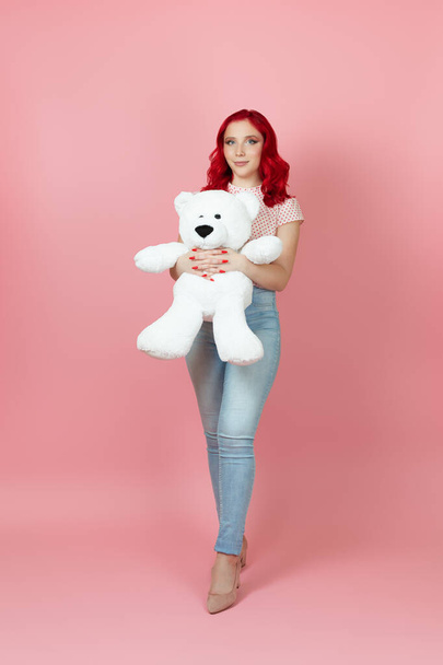 full-length beautiful, delicate woman in jeans with red hair hugs a large white teddy bear isolated on a pink background - Foto, imagen