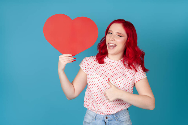 close-up smiling young woman with red hair holds red paper heart, winks, putting out tongue and gives thumbs up isolated on blue background - Foto, afbeelding