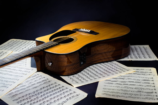 Notenblätter mit Partituren und akustischer klassischer gelber sechssaitiger Gitarre mit schwarzem Pickguard auf isoliertem schwarzem Hintergrund - Foto, Bild