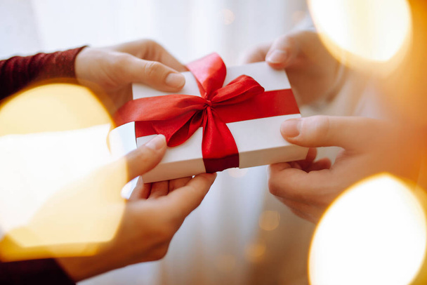Valentine's day concept. Young romantic couple holding a gift in their hands with a garland lights bokeh on the front. White present box with a red ribbon. 14 February celebration together - Fotoğraf, Görsel