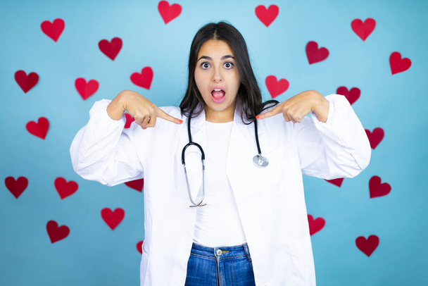 Nuori lääkäri nainen yllään lääketieteellinen takki ja stetoskooppi yli sininen tausta punaiset sydämet etsivät luottavainen hymy kasvoilla, osoittaa itseään sormilla ylpeä ja onnellinen. - Valokuva, kuva