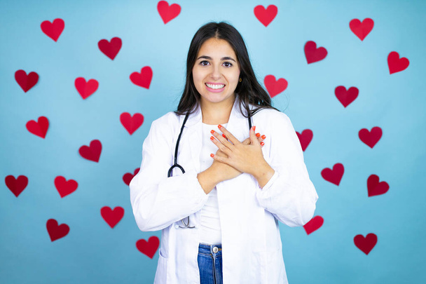 Young doctor woman wearing medical coat and stethoscope over blue background with red hearts smiling with her hands on her chest and grateful gesture on her face. - Valokuva, kuva
