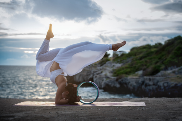 Mujer joven ejercita yoga en la naturaleza vista al atardecer del mar. Poses de Yoga Modificados con Rueda de Yoga, Estiramiento y Fortalecimiento de Ejercicios con Yoga Avanzado, Relajación y Concepto de Cuidado de la Salud. - Foto, imagen