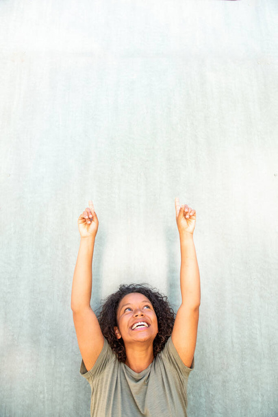 Портрет весела молода афроамериканка з піднятими руками і пальцями, що вказують на простір для копіювання. - Фото, зображення