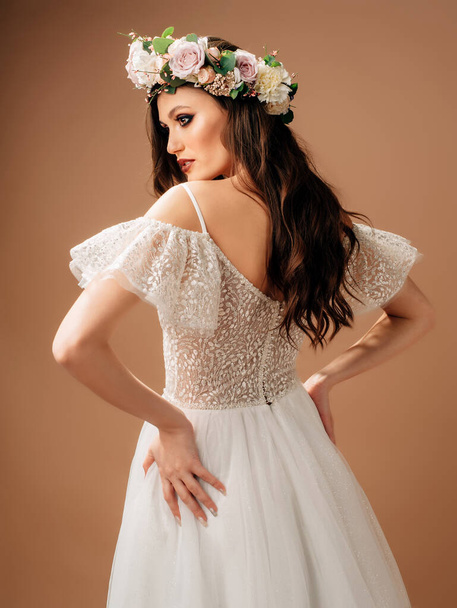 Happy bride in lush wedding dress posing in studio on brown background. Spring posh wedding dress with long lace sleeves and floral garland. - 写真・画像