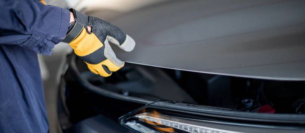 Mies auto mekaanikko käsi yllään keltainen käsine avaaminen auton konepelti. Autojen osien ja moottoreiden tarkastaminen autoteollisuudessa. Auton huolto ja korjaus käsite - Valokuva, kuva