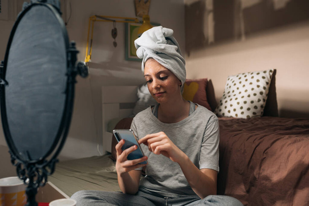 girl applying cosmetic face mask, sitting in her room , using mobile phone - Foto, afbeelding