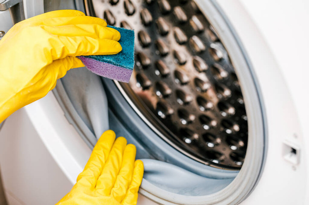 Ένας άντρας με κίτρινα γάντια καθαρίζει ένα βρώμικο, μουχλιασμένο λάστιχο σε ένα πλυντήριο. Μουλάρι, χώμα, λέπια στο πλυντήριο. Περιοδική συντήρηση οικιακών συσκευών. - Φωτογραφία, εικόνα