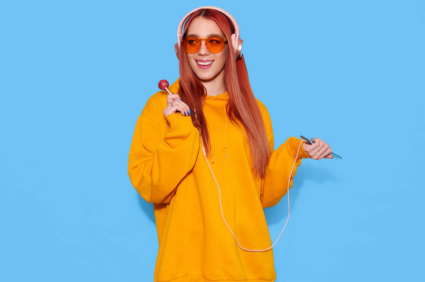 Хіпстерська дівчина-підліток у модному спокійному светрі їсть льодяник і слухає музику в навушниках на синьому фоні
 - Фото, зображення