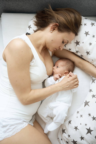 Beautiful woman breastfeeding her baby in a cozy house on the bed. - Фото, изображение
