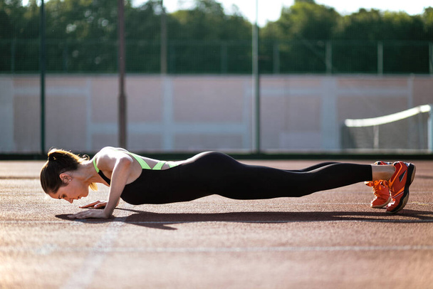 A sporty woman does push-UPS to warm up before an evening workout at the city stadium. Runner training outdoors, the concept of a healthy lifestyle - Foto, immagini