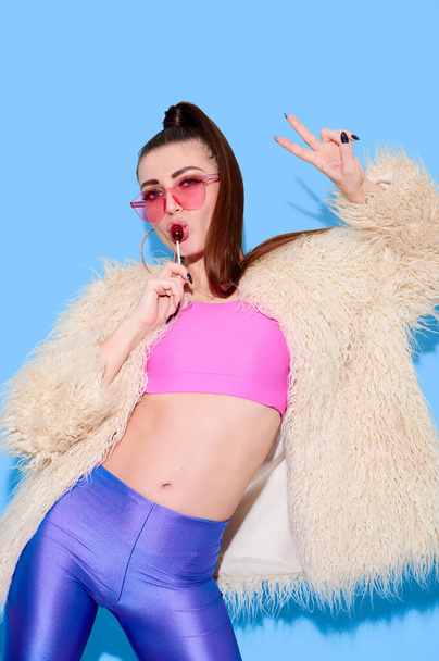 Hipster girl wearing bright colored clothes and fur coat lick lollipop and show victory gesture on blue background - Fotó, kép