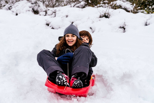 dos niños caucásicos muy felices jugando en la nieve en una montaña con un trineo rojo - Foto, Imagen