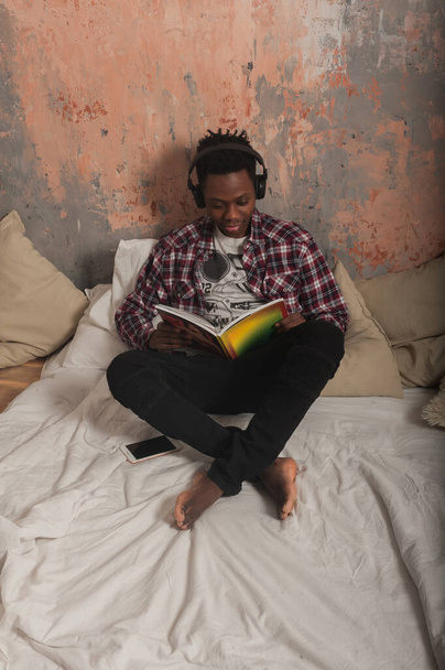 a young african american man lies on the bed wearing headphones and reads a magazine. High quality photo - Φωτογραφία, εικόνα