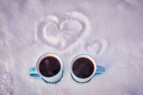 two blue mugs of steaming coffee in the snow with painted hearts in winter park. - Фото, изображение