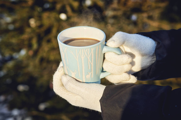 female hands in white gloves with a blue mug of steaming coffee on the background of a winter forest - Foto, afbeelding