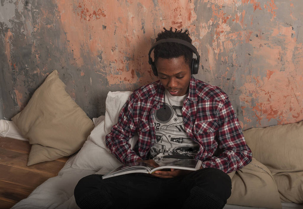 Ένας νεαρός Αφροαμερικάνος ξαπλώνει στο κρεβάτι φορώντας ακουστικά και διαβάζει ένα περιοδικό. Υψηλής ποιότητας φωτογραφία - Φωτογραφία, εικόνα