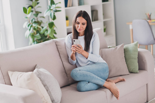 Full body photo of calm charming lovely brunette young woman sit couch wear jeans use phone inside home indoors - Фото, изображение