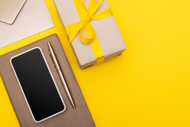 top view of smartphone with blank screen on copy book with pen near envelope and gift box isolated on yellow - Photo, Image