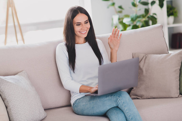 Photo of charming positive woman wave hand say hello videocall on laptop sit sofa home indoors in living room - Fotoğraf, Görsel