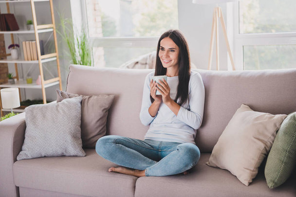 Photo of charming brown haired happy woman hold mug drink tea sit sofa in living room good mood inside indoors - Foto, afbeelding