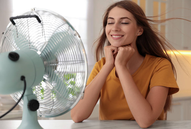 Woman enjoying air flow from fan at table in kitchen. Summer heat - Фото, изображение