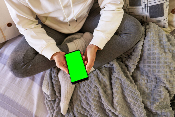 Κάτοψη της νεαρής γυναίκας που κάθεται στον καναπέ φορώντας άνετα ρούχα με το smartphone στο χέρι. Γυναίκα που κάθεται με σταυρωμένα πόδια κρατά το κινητό τηλέφωνο με λευκή πράσινη οθόνη για αντίγραφο χώρου - Φωτογραφία, εικόνα