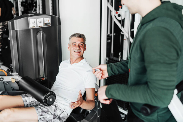 Training of the senior man in a sports hall under the supervision of the rehabilitologist. The man listens carefully to the recommendations of the coach. - Foto, imagen