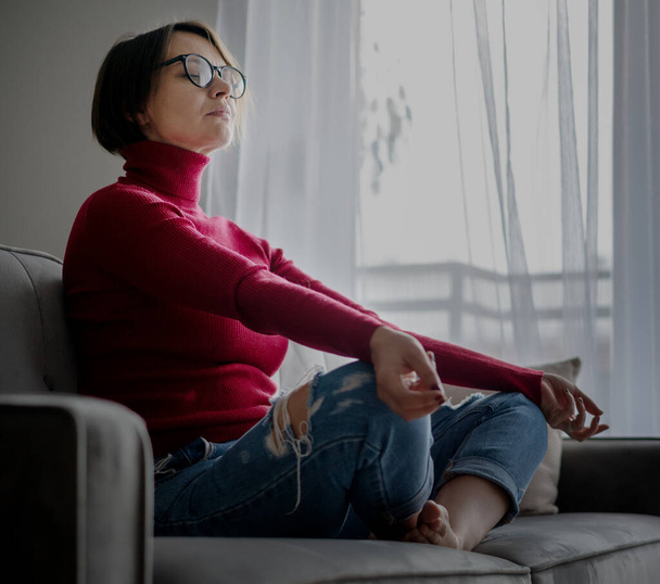 Beautiful mature woman in glasses sitting at home on the sofa meditating in the lotus position on the sofa, taking a break from work, mindfulness and mental health - Fotoğraf, Görsel
