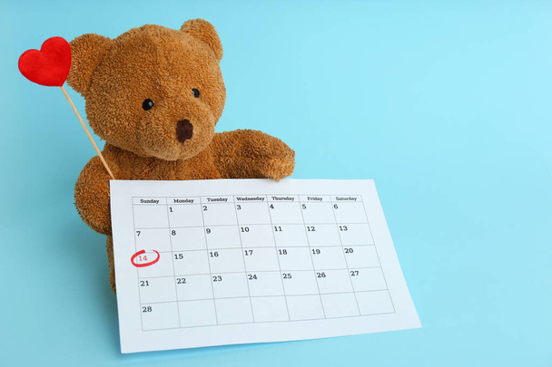 Cute teddy bear with calendar and red heart on light blue background. Valentine's day celebration - Photo, Image