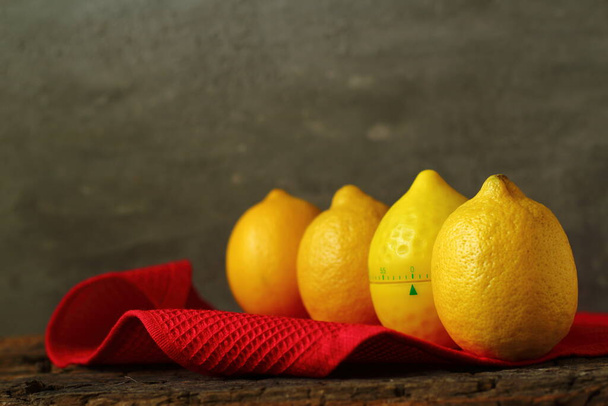 Лимоны фрукты и таймер в виде лимона находятся на старом деревянном столе с красной салфеткой на бетонном фоне, избирательный фокус. - Фото, изображение