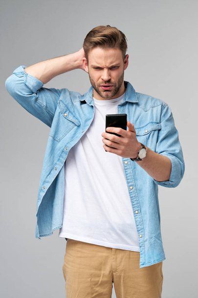 Young shocked stressed angry and frustrated man using smartphone over grey background - Фото, зображення