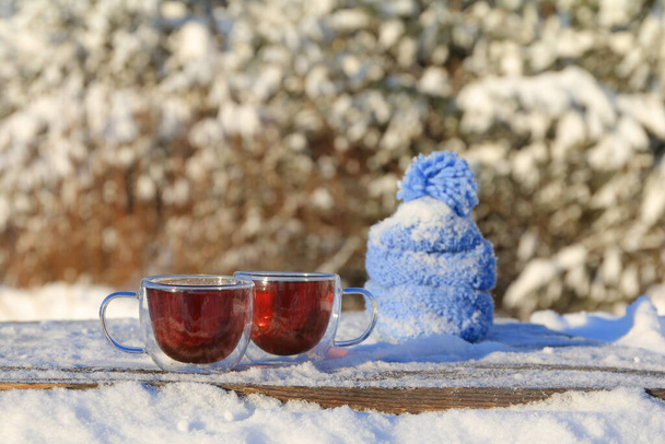 Две стеклянные кружки чая рядом с голубой вязаной шляпой и шарфом стоят на деревянном столе на фоне зимнего пейзажа.   - Фото, изображение